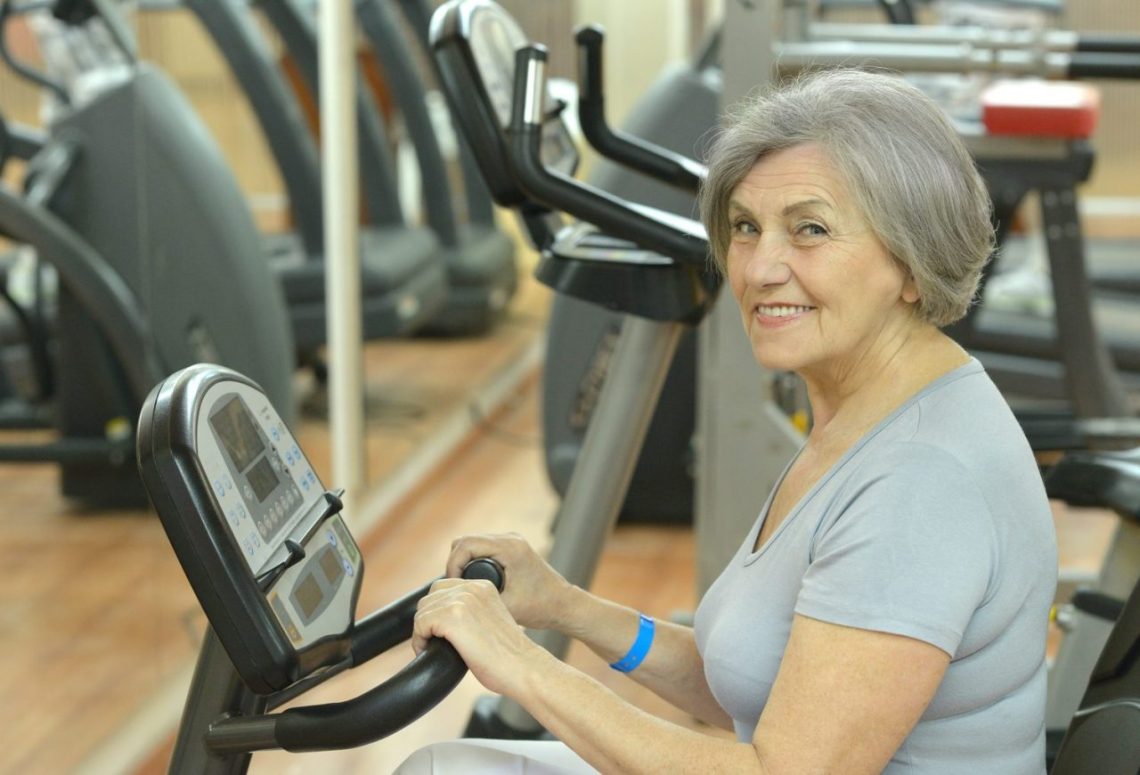 Une femme senior pédale doucement sur un vélo d’appartement pour reprendre l’activité physique après une prothèse du genou.