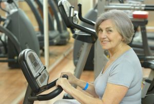 Une femme senior pédale doucement sur un vélo d’appartement pour reprendre l’activité physique après une prothèse du genou.