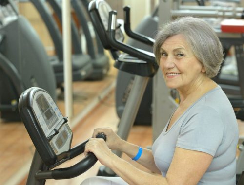 Une femme senior pédale doucement sur un vélo d’appartement pour reprendre l’activité physique après une prothèse du genou.
