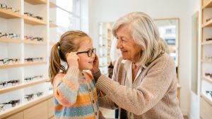 Une grand-mère souriante accompagne sa petite-fille qui essaie des lunettes dans une boutique d’optique lumineuse.