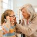 Une grand-mère souriante accompagne sa petite-fille qui essaie des lunettes dans une boutique d’optique lumineuse.