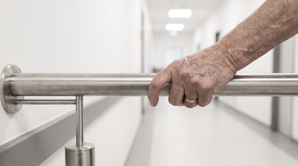 Une personne âgée marche prudemment dans un couloir dégagé équipé d’une rampe murale.